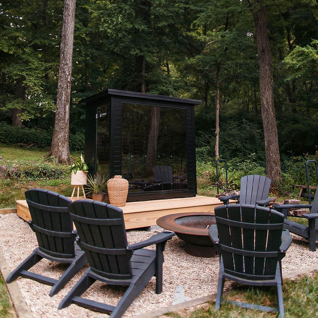 Outdoor seating area with black Adirondack chairs around a fire pit in a forested setting.