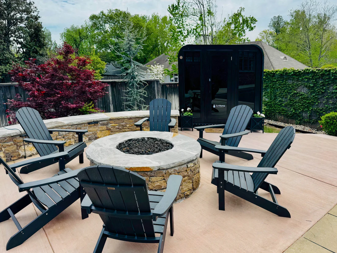 Outdoor patio with stone fire pit and Adirondack chairs surrounded by greenery.