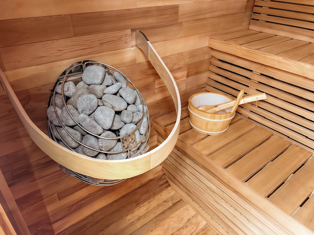 Close-up of a sauna heater with stones and wooden bucket inside a wooden sauna.